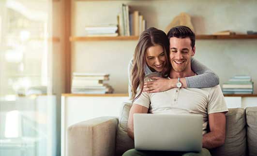 A man and woman sitting on the couch looking at a laptop.